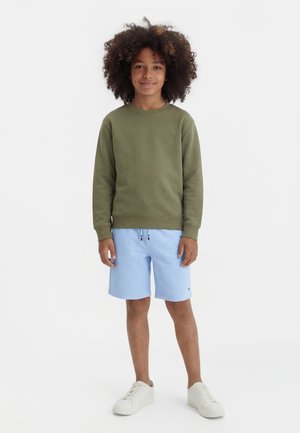 Αγόρι με σγουρά μαλλιά που φοράει φούτερ σε ελιά πράσινο χρώμα, ανοιχτό μπλε σορτς με κορδόνια και λευκά αθλητικά παπούτσια, στέκεται και χαμογελάει.