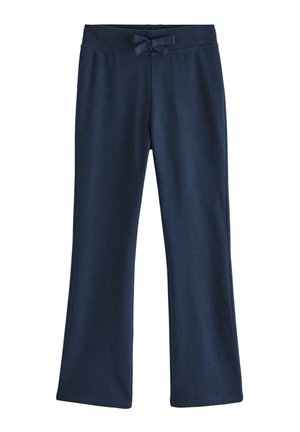 Pige med langt brunt hår iført navyblå pelskantet jakke, blomstret top, navy bukser og beige sko, står med hænderne bag ryggen.