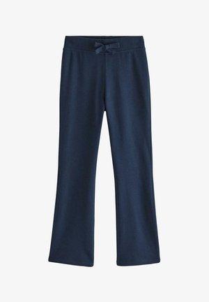 Pige med langt brunt hår iført navyblå pelskantet jakke, blomstret top, navy bukser og beige sko, står med hænderne bag ryggen.