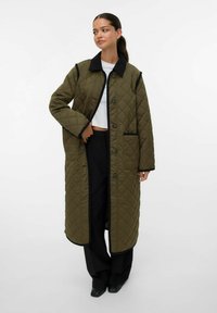 Kvinde iført en lang olivengrøn quiltet frakke med sort kant, hvid cropped top, sorte bukser med brede ben og sorte sko, stående foran en hvid baggrund.