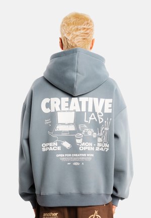 Osoba z krótkimi blond włosami, nosząca niebiesko-szarą bluzę z kapturem z białym tekstem "CREATIVE LAB" i ilustracjami przestrzeni roboczej na plecach.