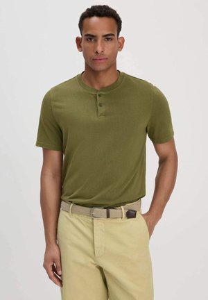 Junger Mann, der ein olivgrünes Henley-Shirt mit kurzen Ärmeln und eine beige Hose mit einem geflochtenen Gürtel trägt, vor einem einfarbigen hellen Hintergrund steht.