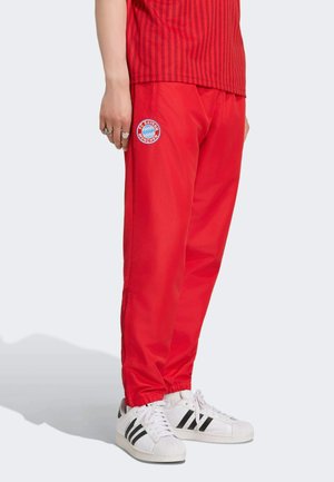 Osoba nosząca czerwone spodnie FC Bayern München z elastycznymi mankietami i białe sneakersy Adidas z czarnymi paskami, stojąca bokiem.