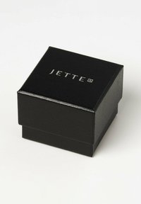 Sort smykkeboks med tekstureret overflade, med et sølvpræget "JETTE" logo på toppen. Kompakt design uden yderligere detaljer.