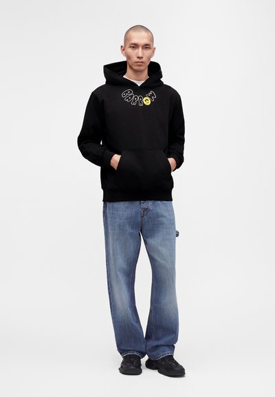Homme portant un sweat à capuche noir avec le texte "Parron" et un visage souriant, jean bleu ample et baskets noires, debout les mains dans la poche du sweat.
