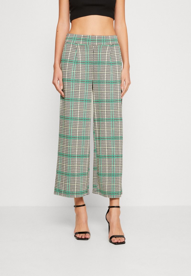 Femme portant un pantalon large taille haute, court, à motif écossais vert et noir, avec des sandales à talons hauts noires à brides.