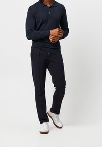 Marineblaues langärmliges Polohemd mit dreiknöpfiger Knopfleiste, kombiniert mit dunklen Hosen und weißen Sneakers mit braunen Akzenten.