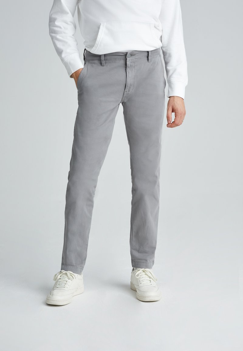 Homme portant un pantalon slim gris clair, un sweat-shirt blanc et des baskets blanches, debout avec une main dans la poche sur un fond uni.