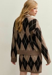 Femme blonde portant un pull en argyle marron et noir et une jupe assortie, se tenant de côté devant un fond de mur clair.