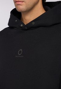 Černá mikina s kapucí vyrobena z měkké tkaniny. Obsahuje stříbrné patentky a diskrétní logo vytištěné na hrudi.