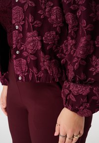 Bordeaux blouse met een getextureerd bloemenpatroon, voorzien van een knoopsluiting aan de voorkant en gerimpelde mouwen, gecombineerd met effen bordeaux nauwsluitende broek.