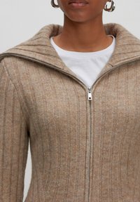 Brauner gerippter Zip-Pullover mit hohem Kragen, ausgestattet mit einem silbernen Reißverschluss und einer strukturierten Oberfläche, darüber getragen mit einem weißen Hemd.