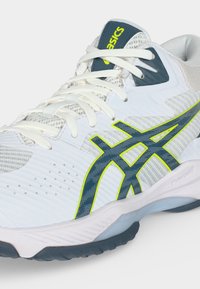 Λευκό και γκρι αθλητικό παπούτσι Asics με σκούρο μπλε λογότυπο με κίτρινο περίγραμμα και λευκά κορδόνια σε ανοιχτό φόντο.