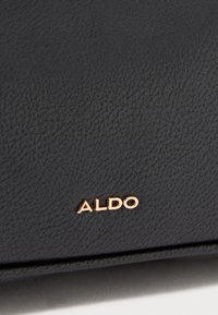 Sac en simili cuir noir texturé avec un logo "ALDO" en rose doré à l'avant. Présente une finition lisse et un design minimaliste.