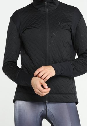 Frau trägt eine schwarze gesteppte Jacke mit Reißverschluss und graue Radlerhose, sie justiert ihr linkes Handgelenk mit der rechten Hand.