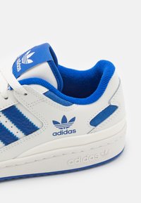 Adidași albi cu dungi din piele albastră, limbă din material textil albastru și talpă din cauciuc texturată. Detalii includ logo-ul și accente albastre.