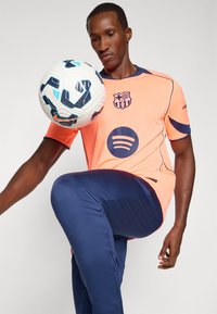 Voetballer in een oranje shirt met blauwe accenten, voorzien van een logo en Spotify branding; marineblauwe broek, die een gemusterde bal controleert.