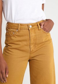 Vaqueros de tiro alto color mostaza con pierna recta, que cuentan con bolsillos delanteros y detalles de costura visibles. Llevados con una camisa blanca básica.