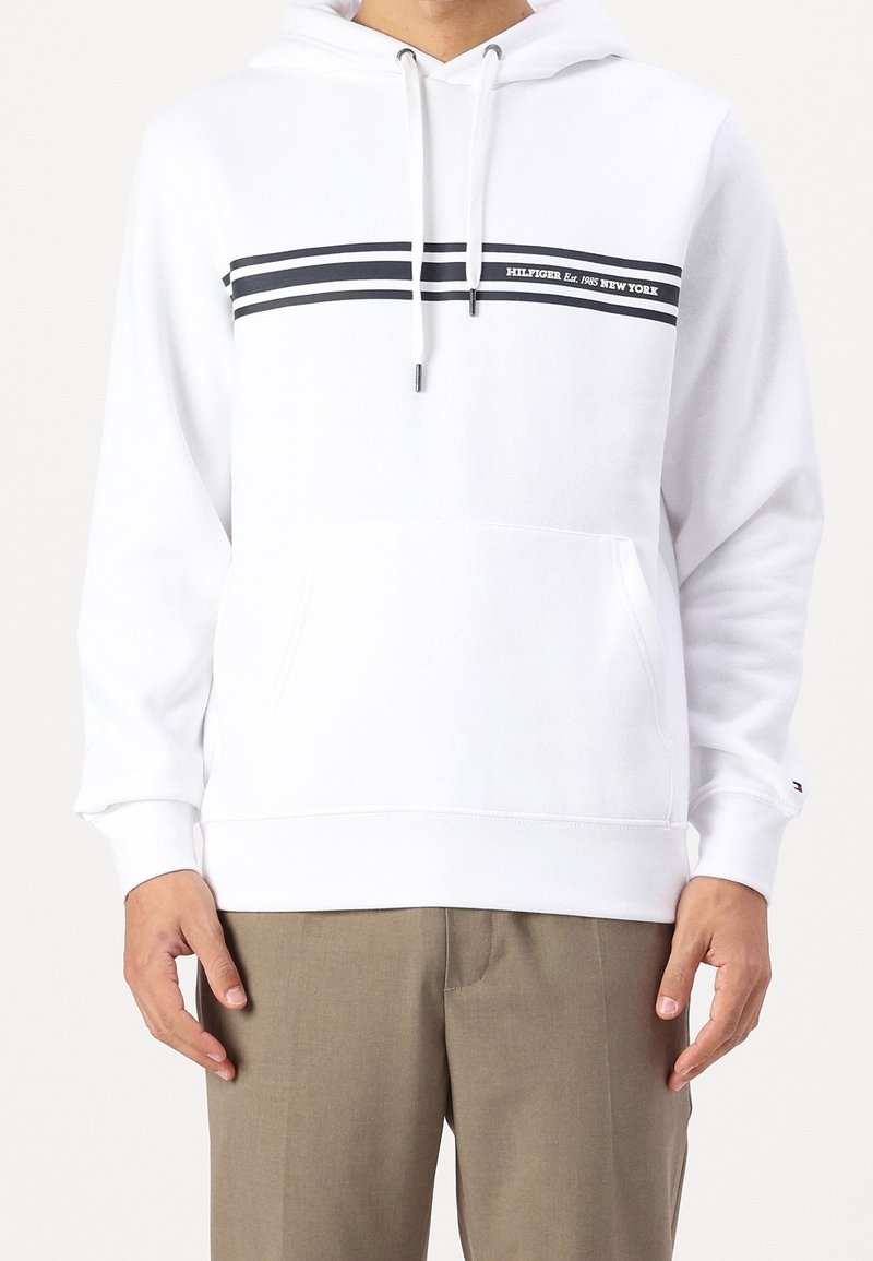 Biała bluza z kapturem z przednią kieszenią kangurka, szarymi troczkami i granatowymi paskami. Na piersi znajduje się logo z tekstem. Miękki bawełniany materiał.