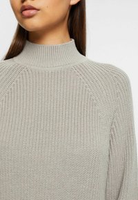 Maglione grigio lavorato a maglia con colletto alto, texture a coste e maniche raglan. Focus sul materiale morbido e sul dettaglio della cucitura uniforme.