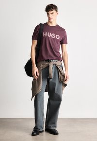 Burgundi T-särk, millel on "HUGO" logo, kombineeritud lõdvade hallide teksadega. Pruun väliskiht on seotud talje ümber. Must õlakott ja mustad kingad.