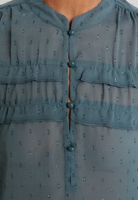Blouse bleue transparente avec petits pois brillants, boutons ronds en tissu et détails à volants horizontaux sur la poitrine et la taille.