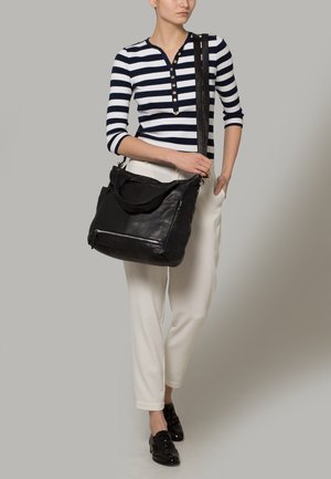 Kobieta w pasiastym topie i białych spodniach, trzymająca dużą czarną skórzaną torbę na ramię z przednią kieszenią na zamek.