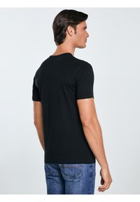 Mann mit dunklen Haaren steht seitlich und trägt ein einfaches schwarzes kurzärmeliges T-Shirt und blaue Jeans vor neutralem Hintergrund.