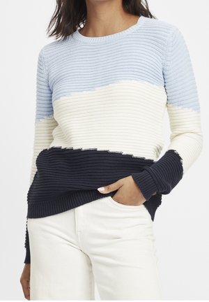 Femme portant un pull en maille côtelée avec des blocs de couleurs horizontaux en bleu clair, blanc et marine, associé à un pantalon blanc, main dans la poche.