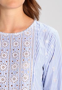 Blouse rayée bleu clair et blanche avec des motifs floraux brodés et des détails en œillets ouverts le long de l'avant et des manches.