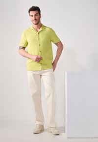 Camisa corta de manga verde claro con tejido texturizado y botones marrones, combinada con pantalones off-white y zapatillas gris claro.
