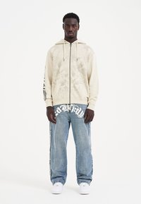 Un hombre está de pie frente a un fondo blanco, vistiendo una sudadera ligera con un diseño gráfico, jeans holgados con letras llamativas y zapatillas blancas.