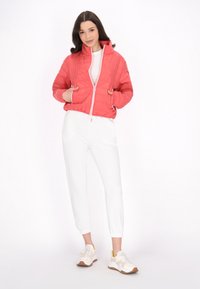 Giacca trapuntata corallo con collo alto e cerniera frontale, abbinata a pantaloni jogger bianchi. Il modello indossa sneaker beige con dettagli gialli.