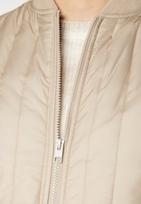 Veste bomber matelassée beige avec un col côtelé, une fermeture éclair argentée et des coutures verticales. Portée par-dessus un pull léger texturé.