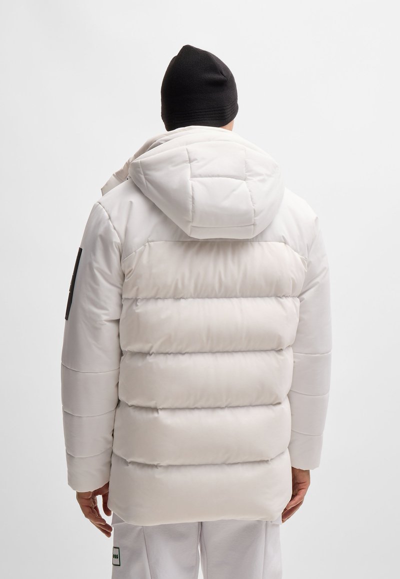 Weiße Puffjacke mit Kapuze, ausgestattet mit genähten horizontalen Abschnitten und einem matten Finish. Die Jacke hat einen schwarzen Patch am linken Ärmel.