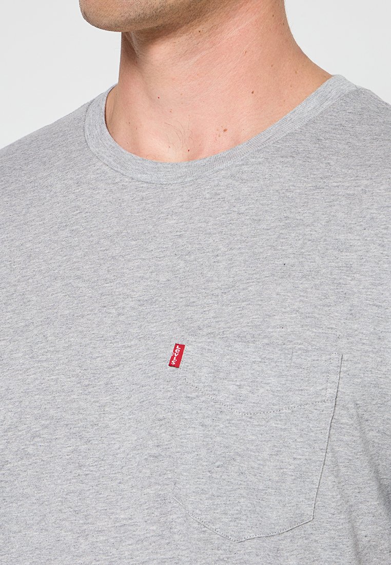 T-shirt en coton gris à col ras du cou avec une poche sur le côté gauche et une étiquette rouge. Texture douce, manches courtes et coupe décontractée.