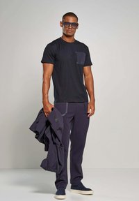 Marineblauwe t-shirt met korte mouwen en een zak, gecombineerd met donkere broek met contrasterende stiksels. Het model houdt een jack vast en draagt blauwe zonnebril.