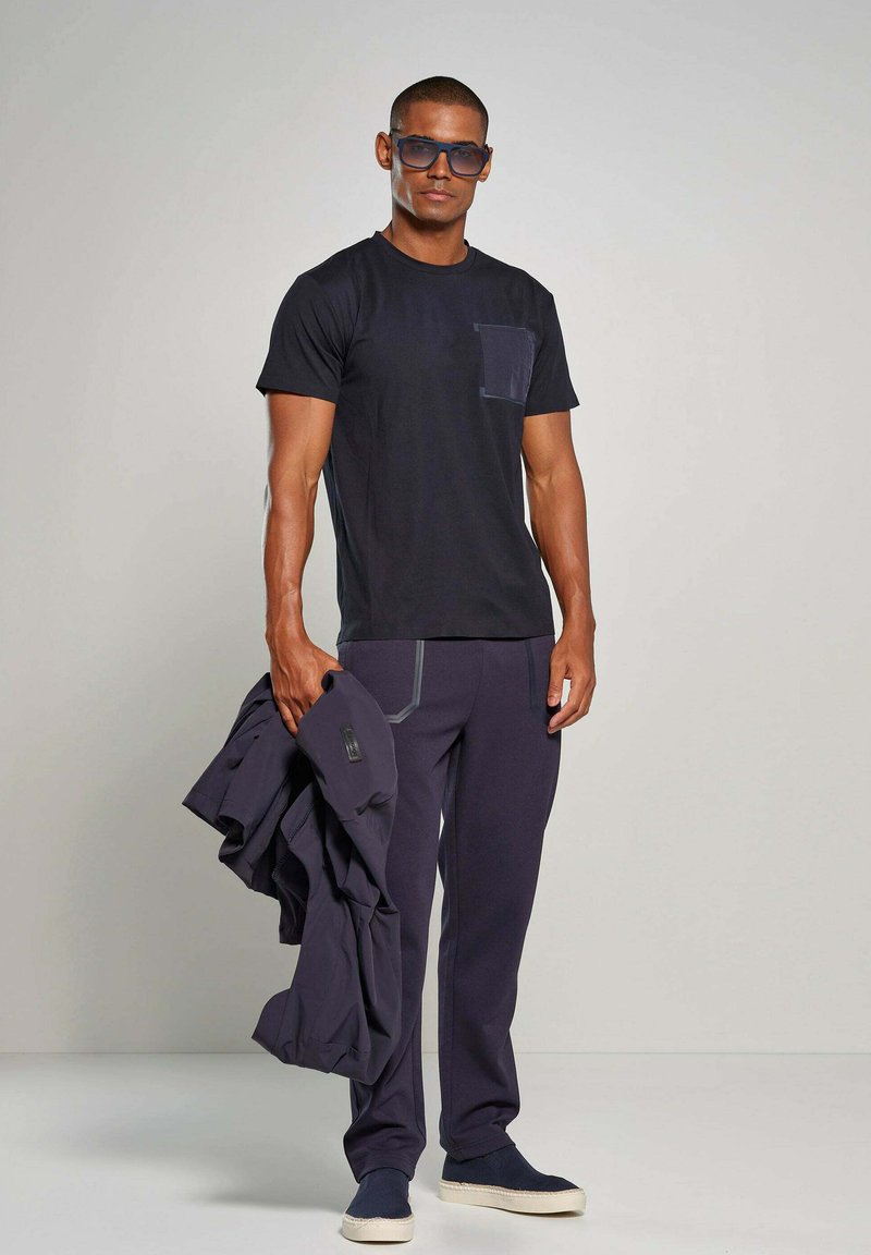 Marineblauwe t-shirt met korte mouwen en een zak, gecombineerd met donkere broek met contrasterende stiksels. Het model houdt een jack vast en draagt blauwe zonnebril.