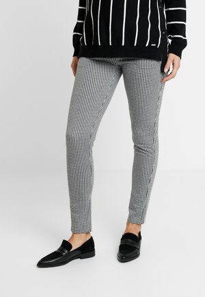 Nogi w czarno-białych spodniach w kratkę w jodełkę, połączone z czarnymi wsuwanymi mokasynami i czarnym swetrem z pionowymi białymi paskami.