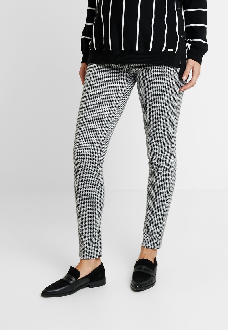Jambes portant un pantalon à motif pied-de-poule noir et blanc, associé à des mocassins noirs sans lacets et un pull noir avec des rayures blanches verticales.