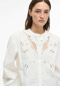 Balta kokvilnas blūze ar izsmalcinātu izšuvumu un izgriezumu detaļām priekšpusē. Tai ir apaļa apkakle un aizdare ar pogām.