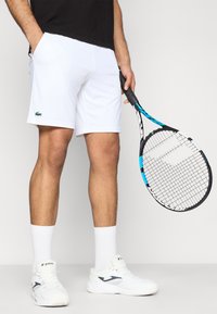 Λευκά σορτς τένις από ελαφρύ ύφασμα, με πλαϊνές τσέπες και πράσινο λογότυπο. Μια μαύρη και μπλε ρακέτα τένις κρατιέται στο ένα χέρι.