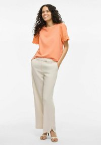 Blusa de color melocotón con mangas con encaje, combinada con pantalones de pierna ancha en un tono beige claro, con aberturas laterales. Las sandalias completan el atuendo.