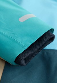 Ärmel einer türkisfarbenen Jacke mit einem gerippten marineblauen Bündchen, glatter Textur und einem reflektierenden Akzent. Nahaufnahme von Stoff- und Nähdetails.
