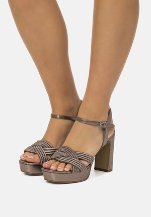 Piedi che indossano sandali con plateau e tacco alto metallici marroni con cinturini intrecciati e chiusure con fibbia alla caviglia su uno sfondo semplice.