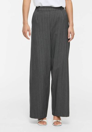 Femme portant un pantalon large à rayures gris foncé taille haute et des sandales blanches à bout ouvert sur fond blanc.