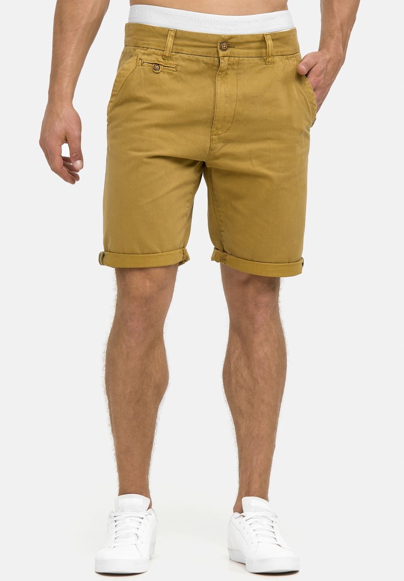 Shorts en coton beige avec revers roulés, poches avant et fermeture à un bouton. Associés à des baskets de sport blanches.