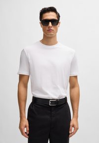 White cotton t-shirt with short sleeves, paired with black trousers and a black belt. The model wears large black sunglasses, facing forward.