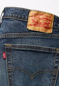 Vidutinio mėlynumo džinsai, su oranžinėmis siūlėmis, odiniu ženkleliu gale ir raudonu Levi's etikete ant kišenės.