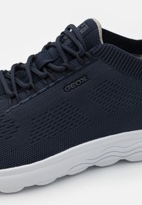 Námořnické modré tenisky s texturovaným, prodyšným svrškem, tmavými tkaničkami a bílou podrážkou. Na boku je logo Geox a perforovaný design pro ventilaci.